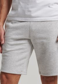 Hombre vistiendo pantalones cortos casuales gris claro y una camiseta blanca, de pie con las manos relajadas a los lados.