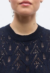 Πλεκτό πουλόβερ σε ναυτικό μπλε χρώμα με υφή ανοιχτού δαντελένιου μοτίβου και ριμπ λαιμόκοψη. Διακοσμημένο με γεωμετρικά σχήματα στο σχέδιο του υφάσματος.