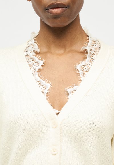 Κοντινό πλάνο ενός ατόμου που φοράει λευκή ζακέτα με δαντέλα σε σχήμα κυμάτων κατά μήκος του V-λαιμού.