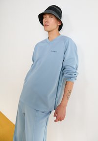 Light blue sweatshirt with a v-neck, long sleeves, and subtle side stripes. Paired with matching pants, displaying the brand logo. Black bucket hat.