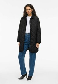 Svart vadderad lång kappa med krage, knäppning och två sidofickor, bärs över en vit topp och blå jeans. Svarta högklackade skor.
