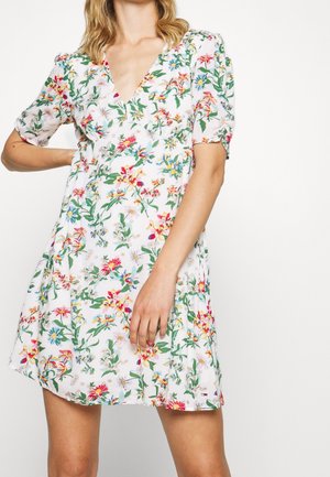 Das Model posiert in einem blumenmusterartigen Kleid mit tiefem V-Ausschnitt, das pinke, blaue und grüne Blumen auf einem weißen Hintergrund zeigt.