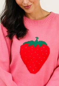 Pull en laine rose fabriqué à partir d'un matériau tricoté doux, mettant en avant un grand motif de fraise rouge avec des feuilles vertes et des motifs de graines blanches.