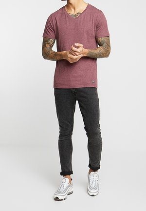 Camiseta de cuello en V color marrón de tejido suave, combinada con jeans oscuros ajustados. Llevada con zapatillas blancas y grises, mostrando un aspecto casual.