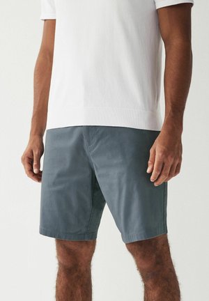 Homme portant une chemise blanche unie à manches courtes et un short gris arrivant aux genoux, debout devant un fond uni.