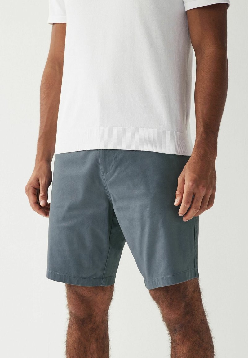Homme portant une chemise blanche unie à manches courtes et un short gris arrivant aux genoux, debout devant un fond uni.