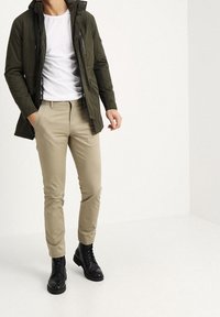 Voděodolná bunda olivově zelené barvy, bílé tričko, béžové kalhoty zúženého střihu a černé šněrovací boty. Jednoduchý design, přiléhavý tvar a hladká textura.