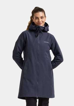 Frau mit hellbraunem Haar im Pferdeschwanz, die einen langen, dunkelblauen Didriksons Regenmantel mit Kapuze und Frontreißverschluss trägt, steht mit den Händen hinter dem Rücken.
