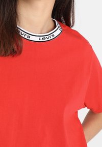 Camiseta de algodón roja con cuello redondo que presenta un collar acanalado en blanco y negro etiquetado como "Levi's". Mangas cortas y ajuste relajado.