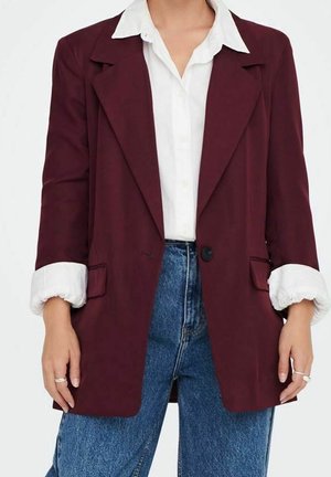 Kvinde iført en hvid skjorte med op-rullede ærmer under en bordeaux blazer og blå jeans med høj talje, hænderne afslappet ved siderne.