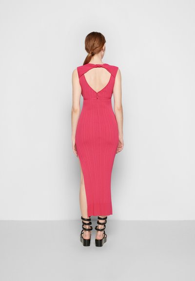 Vestido longo rosa curvilíneo com costas em V profundas, apresentando um design em torção nos ombros e uma fenda lateral. Estilizado com sandálias pretas de tiras.