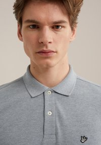 Pelēka polo krekla ar tekstūrētu audumu, divām gaišām pogām un mazu izšūtu grafiku kreisajā pusē. Klasisks apkakles dizains.