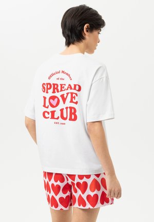 Persoon in wit T-shirt met rode tekst "Spread Love Club" en hartafbeelding, gecombineerd met witte korte broek bedekt met rode harten, rug naar de kijker.
