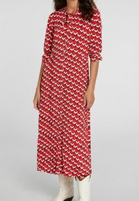 Robe mi-longue rouge avec un motif géométrique blanc, avec un devant à boutons et des manches longues. Portée avec des bottes de cowboy blanches.