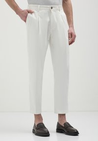 Pantaloni bianchi con una vestibilità su misura e due pieghe frontali, abbinati a mocassini marrone scuro, su uno sfondo semplice.