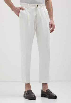 White trousers featuring a tailored cut and two front pleats, paired with dark brown loafers, set against a plain background.