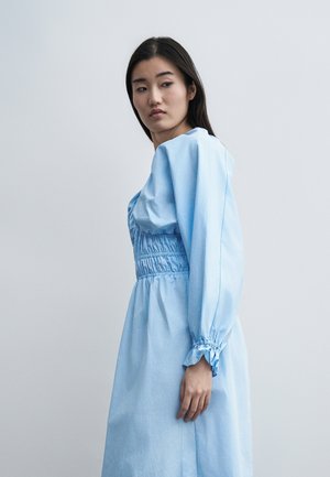 Ung kvinde med langt mørkt hår iført en lyseblå langærmet kjole med indsnøring i taljen og ved ærmerne, står mod en ensfarvet grå baggrund.