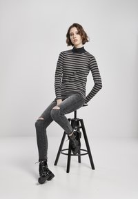 Langærmet sømandsstrik med striber i sort og grå, parret med slidte mørke jeans og sorte laklæder snørebåndsstøvler, siddende på en sort skammel.