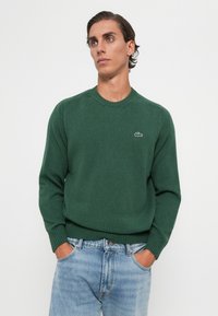 Bărbat îmbrăcat într-un pulover verde închis cu un mic logo de crocodil, blugi albaștri deschiși, cu mâinile în buzunare, privind ușor spre dreapta pe un fundal alb.