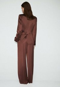 Brauner Satinanzug mit einem maßgeschneiderten Blazer mit Reverskragen und weit geschnittenen Hosen, der eine glatte Textur und ein strukturiertes Design zeigt.