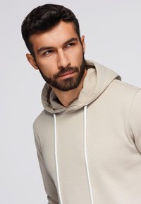 Béžová mikina s měkkou texturou, vybavená stahovací šňůrkou a dvoubarevným designem. Model má pečlivě upravený plnovous a středně dlouhé vlasy.