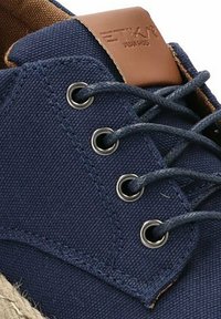 Zapato de lona azul marino con ojales de metal y cordones azules. Acento de cuero marrón en el talón y suela de yute natural. Superficie texturizada.