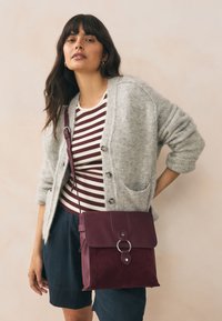 Bordeauxrode leren en suède schoudertas met een voorklep, ronde metalen details en een strak ontwerp. Draagt deze schuin over het lichaam, gecombineerd met een gestreept shirt en een vest.