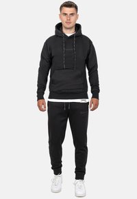 Zwarte hoodie met trekkoord, voorzak en logo. Gepaard met zwarte joggers en witte sneakers, tegen een neutrale achtergrond.