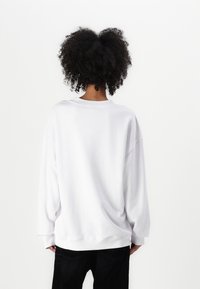 Fehér, oversize kapucnis pulóver, leeső vállakkal, bordázott nyakkal és mandzsettákkal, sima textúrával, sötét farmernadrággal viselve, hátulról nézve.