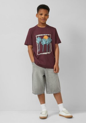 Jongen staat met handen in de zakken, draagt een bordeauxrood T-shirt met palmboomprint, grijze knielange shorts, witte sokken en witte sneakers.