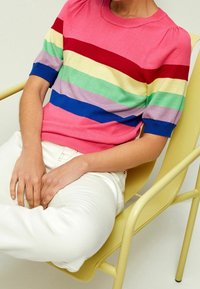 Kortærmet strikket sweater i pink med vandrette striber i rød, gul, grøn, blå og lilla. Båret med hvide bukser, siddende på en gul stol.