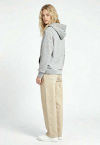 Donna con capelli biondi lunghi e ricci, indossa una felpa grigia con cappuccio, pantaloni beige a gamba larga e scarpe da ginnastica bianche, in piedi davanti a uno sfondo chiaro uniforme.