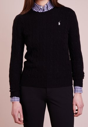 Osoba ubrana w czarny sweter z grubym splotem z białym logo, na niebiesko-białej kraciastej koszuli, w połączeniu z czarnymi spodniami na jednolitym tle.