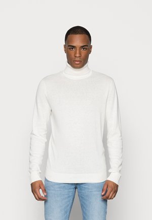 Suéter blanco de cuello alto hecho de tela ligera, con mangas largas y puños y dobladillo acanalados, combinado con unos jeans de color azul claro.