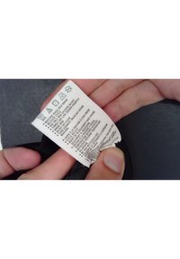 Označení péče na látce, bílé s černým potiskem, obsahující pokyny pro praní a údržbu. Ruka držící štítek na tmavém pozadí.