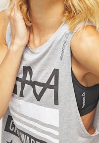 Canottiera grigia senza maniche con grandi lettere grafiche nere e strisce orizzontali. Presenta un ampio scollo e un look a strati sopra un reggiseno sportivo nero.