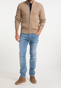 Cárdigan beige de canalé con cremallera y cuello alto, combinado con jeans ajustados azul claro y zapatos de ante marrones.