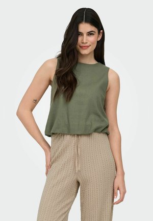 Joven con cabello largo y oscuro, vistiendo una blusa sin mangas de color verde oliva y pantalones beige texturizados, de pie con una mano en la cadera.