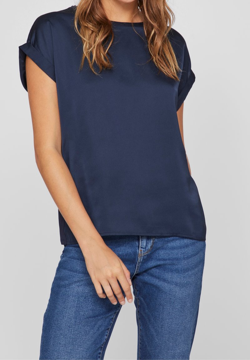 Femme portant un chemisier bleu marine à manches courtes retroussées et un jean en denim bleu, debout devant un fond clair uni.