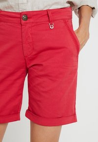Pantalones cortos de algodón rojos con dobladillo enrollado, que presentan un cierre de botón y gancho, bolsillos laterales y costuras decorativas en la cinturilla.