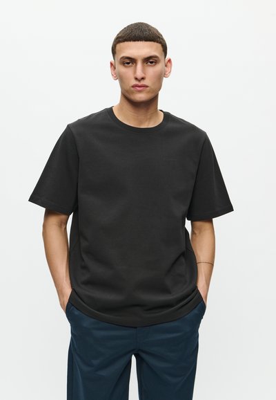 Zwarte t-shirt met korte mouwen van katoen met een ronde hals; heeft een subtiele logo op de borst; gecombineerd met donkerblauwe broeken.