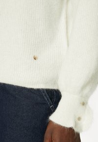 Bílý pletený svetr s jemnou texturou. Má nadýchané rukávy, dva knoflíky na manžetě a malý zlatý detail s logem blízko spodního okraje.