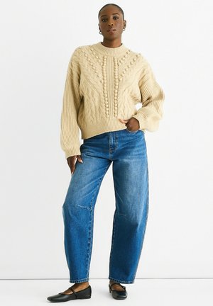 Beige cable-knit sweater with textured patterns, round neckline, and long sleeves paired with blue straight-leg jeans and black flats.