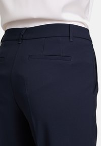 Pantaloni tailored blu navy con una texture liscia, caratterizzati da una parte frontale piatta e due tasche posteriori. La vita è elegante e minimalista.