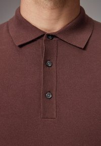 Braunes Strick-Poloshirt mit Kragen, ausgestattet mit einer Knopfleiste mit vier schwarzen Knöpfen und einer strukturierten Stoffoberfläche.