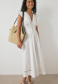 Robe blanche, longueur genou, avec un décolleté en V profond, taille froncée et bordure en œillets à l'ourlet. Associée à un sac en paille tissé et des sandales.