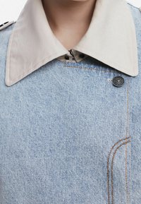 Kurtka dżinsowa z jasnobeżowym kołnierzem, ciemnym zapięciem na guziki i pomarańczowym przeszyciem. Przeszły tekstura i dopasowany krój.