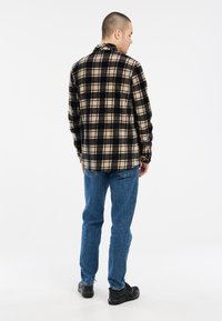 Camisa de franela a cuadros en negro y beige, de manga larga, con un corte relajado, combinada con jeans azules y zapatos negros. Fondo blanco liso.