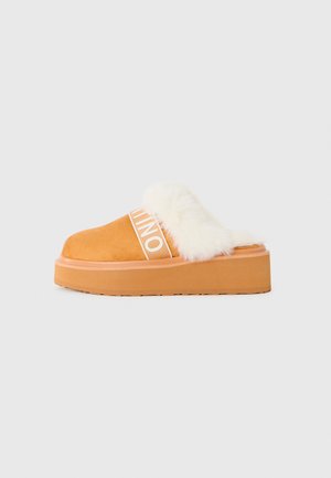 Zapato de plataforma marrón sin cordones con forro de piel de oveja blanca y tira elástica de marca visible sobre un fondo blanco.