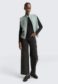 Gilet en maille vert clair avec col montant et devant ouvert, porté sur un top noir à manches longues et associé à un pantalon large foncé et des chaussures noires.